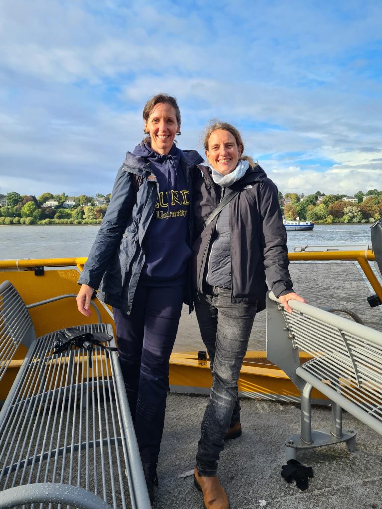 To people standing in the sun on a ferry looking in to the camera and smiling. Photo.