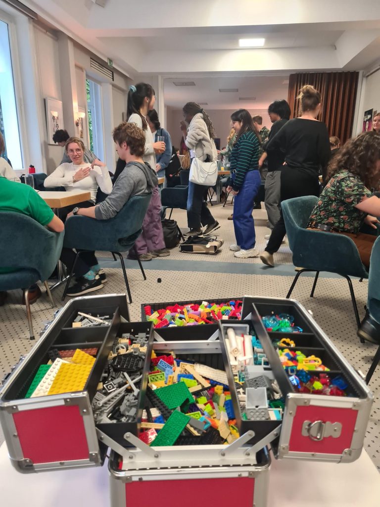 A chest filled with lego, in the background people are standing and sitting by tables. Photo.